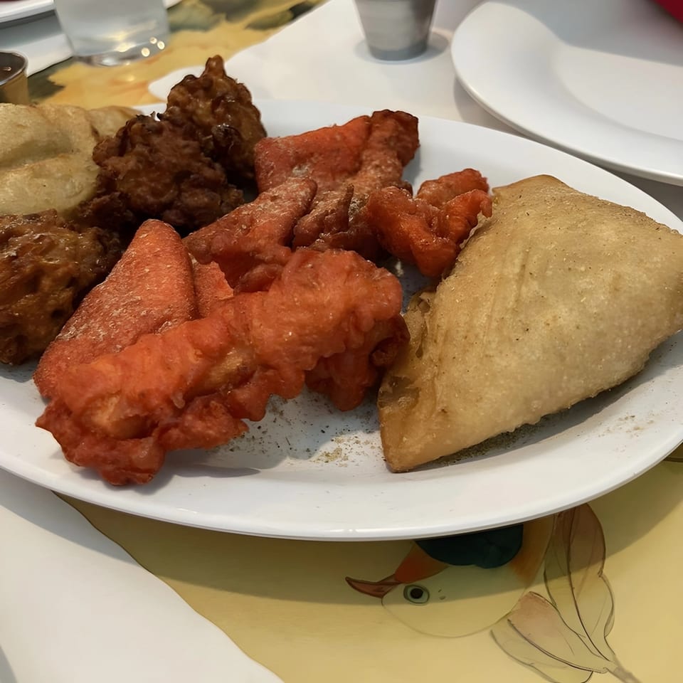 A white plate holds various Indian appetizers, including red fried chicken, brown pakoras, and a samosa.